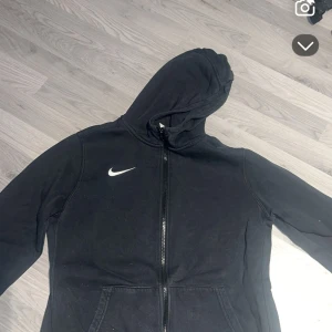 Svart Nike hoodie med dragkedja XL - Svart hoodie från Nike i storlek XL med vit logga på bröstet. Tröjan har huva, dragkedja framtill och två fickor. Perfekt för chill dagar eller när du vill ha en sportig look. Tillverkad i mjukt material för extra komfort. Den passar endast folk som är runt 158-164