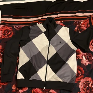 Argyle tröja från Fred Perry M - Tröja från Fred Perry i svart, grått och vitt med stort argylemönster framtill. Tröjan har dragkedja, ståkrage och ribbade muddar. Tillverkad i polyester och har två fickor på sidorna. 