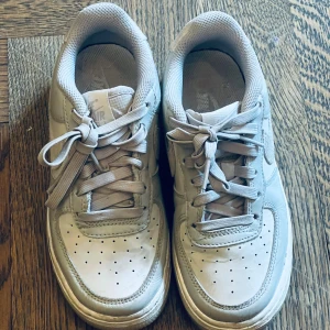 Nike Air Force 1 vita sneakers - Klassiska Nike Air Force 1 sneakers i helvitt skinn med matchande vita snören och perforerad tå. Swoosh-loggan och detaljer i mocka ger en snygg kontrast. Lågt skaft och platt sula för en clean och tidlös look.