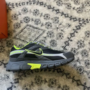 Nike sneakers svart/grå med neon - Nike sneakers i svart och grått med detaljer i neon-gult och silver. Skorna har mesh och syntetpaneler, grå snörning och en robust sula med neoninslag. Swoosh-loggan är markerad med neon och insidan är fodrad i samma färg. Perfekta för dig som gillar sportig och modern stil.