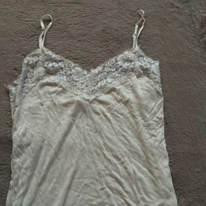 Beige linnetopp med spets från ONLY - Supersöt beige linnetopp från ONLY med tunna axelband och v-ringad spetskant upptill. Toppen har ribbad struktur och är i ett mjukt, lätt material som känns skönt mot huden. Perfekt att styla lager på lager eller som den är under varma dagar.