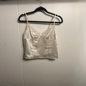 Champagnefärgad satinlinne från H&M Divided - Champagnefärgad linne från H&M Divided i satinliknande polyester med tunna axelband och spetsdetaljer längs v-ringningen. Croppad modell med mjuk glans och liten rosett framtill. Perfekt för att ge en lyxig touch till din outfit.