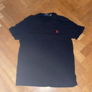 Svart t-shirt Polo Ralph Lauren - Svart t-shirt från Polo Ralph Lauren i mjuk bomull. Klassisk passform med korta ärmar och rund hals. På bröstet finns den ikoniska röda broderade Polo-loggan. Perfekt basic med stilren vibe och diskret branding.