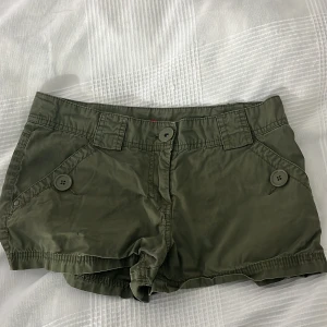Olivgröna shorts med knappar, strl 36 - Snygga olivgröna shorts med flera fickor och dekorativa knappar både fram och bak. Modellen har midwaist och är tillverkad i ett mjukt, lätt polyestertyg som passar perfekt till varma dagar. Stilsäkra detaljer som breda hällor och knappstängning framtill. Liten i storlek och inte så stretchig. Inte avänt alls många tillfällen.