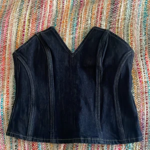 Svart jeanskorsett - Snygg svart korsett i jeansmaterial med en cool v-ringad design. Den har en tight passform och dragkedja bak för enkel på- och avtagning. Perfekt för en festlig kväll eller en utekväll med vänner!