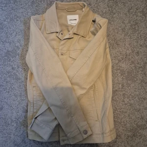 Beige jeansjacka från Jack & Jones - Snygg beige jeansjacka från Jack & Jones i klassisk modell med knappar framtill, två bröstfickor med lock och två sidofickor. Jackan har en rak passform och är tillverkad i bomull. Perfekt för dig som vill ha en stilren och enkel look. Använd en gång!