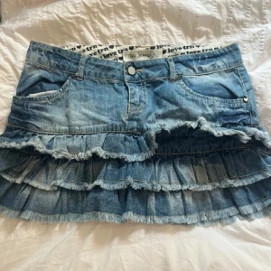 Volangig jeanskjol från PH Jeans - Snygg blå jeanskjol med flera volanglager och fransiga kanter. Klassisk femficksmodell med knapp och dragkedja framtill. Perfekt för dig som gillar en lekfull och trendig stil. Innerlinning med textdetaljer.