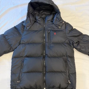 Svart pufferjacka Polo Ralph Lauren - Svart pufferjacka från Polo Ralph Lauren med huva och dragkedja framtill. Jackan har tre fickor med dragkedja och röd logga bröstet. Den är som ny använde den bara förra vintern men den är för stor för mig och därför väljer jag att sälja den. Jackan har inga hål eller repor och ser helt ny ut mvh dennis