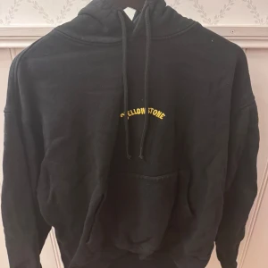 Svart Yellowstone hoodie med tryck - Svart hoodie med huva och snörning, stor färgglad Yellowstone National Park-tryck på ryggen och mindre gul text framtill. Mjuk insida och klassisk känguruficka. Storleken är egentligen L men känns mer som M. Perfekt för dig som gillar coola tryck och naturvibbar. 