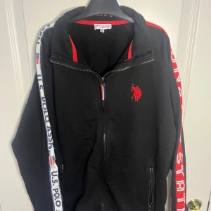 Svart ziptröja U.S. Polo Assn med röd logga - Svart långärmad ziptröja från U.S. Polo Assn med röd broderad logga på bröstet och röda detaljer. Snygga vita och röda band med text längs ärmarna. Hög krage med PLAY-tryck och dragkedjor på fickorna. Perfekt för en sportig och chill stil. Storlek: L 