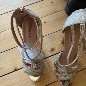 Glittriga guldiga klackar från Perfect - Snygga guldiga klackskor med glittriga remmar och metallic-detaljer. Skorna har öppen tå, hög klack och dragkedja vid hälen. Perfekta för dig som vill sticka ut med extra bling och glam på fötterna.