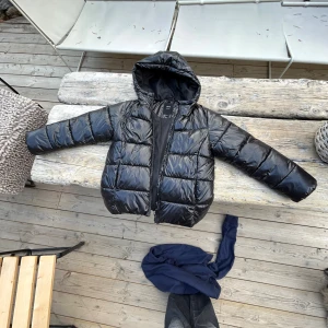 Svart pufferjacka från LMTD XS - Säljer en svart pufferjacka från LMTD i storlek XS. Jackan har glansig finish, huva och dragkedja framtill. Perfekt för kalla dagar med sin vadderade look och klassiska stil. Passar dig som gillar en enkel men snygg vibe.