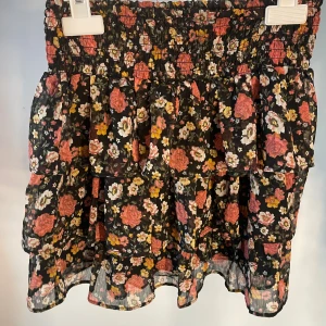 Blommig volangkjol från Lindex - Supersöt kort volangkjol från Lindex med svart botten och blommigt mönster i rosa, gult och vitt. Kjolen har flera lager volanger och resår i midjan för en bekväm passform. Perfekt för dig som gillar en lekfull och färgglad stil. Använd 1-2 gånger