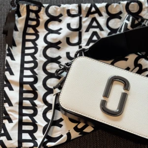 Svart&vit axelväska Marc Jacobs - Säljer en otroligt stilfull axelväska från Marc Jacobs i vit och svart och är perfekt om du inte behöver ha med dig så mycket, för vem orkar att släpa på en stor väska hela tiden? Väskan har två dragkedjor, en bred justerbar axelrem med grafiskt mönster och en praktisk ficka på baksidan. Väskan är köpt på Nelly för ca 2 år sedan och är knappt använd. Pris kan diskuteras…