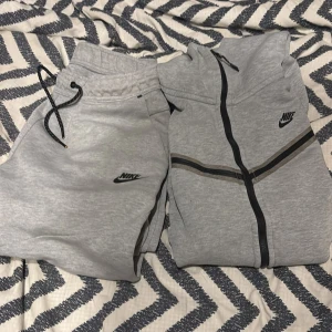 Grå nike tech - Säljer en grå mjukisdress från Nike med matchande hoodie och byxor. Hoodien har svart dragkedja och svarta detaljer, samt Nike-logga på bröstet. Byxorna har snörning i midjan och Nike-logga på benet. Perfekt för chill eller träning.