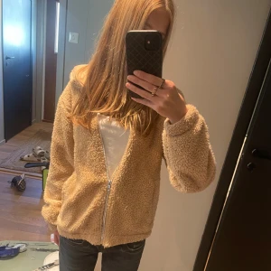 Beige teddyjacka med huva och dragkedja - Mysig teddyjacka i beige med huva och hel dragkedja framtill. Jackan har en loose passform och är tillverkad i fluffigt fleece-material som ger en skön och chill vibe. Perfekt för dig som gillar streetstyle och vill ha något varmt och snyggt.