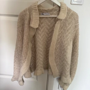 Beige stickad kofta från Zara - Mysig beige stickad kofta från Zara med snyggt sicksackmönster. Koftan har bred ribbad krage och muddar vid ärmslut och nederkant. 