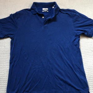 Blå pikétröja från Gant i bomull - Säljer en klassisk blå pikétröja från Gant i 100% bomull. Tröjan har korta ärmar, krage med två knappar och diskret broderad logga på bröstet. Perfekt för en stilren och avslappnad look.