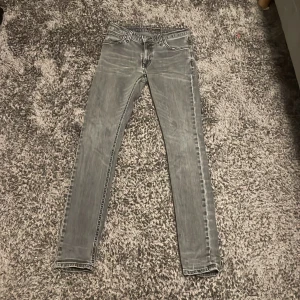 Grå slim jeans från Nudie Jeans W29 L32 - Säljer ett par grå slim jeans från Nudie Jeans med slitna detaljer och klassisk femficksdesign. Jeansen har en smal passform och är tillverkade i mjukt denimtyg. Snygga kontrastsömmar och svart logopatch baktill ger extra edge.