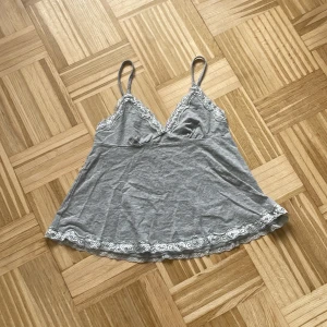 Grå linnetopp med spets från Romwe - Supersöt grå linnetopp från Romwe i storlek XS. Toppen har tunna axelband och är dekorerad med vit spets längs v-ringningen och nederkanten. Perfekt för lager-på-lager eller som statement-piece till jeans eller kjol.
