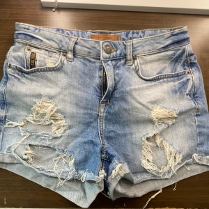 Jeansshorts  - Säljer ett par blå jeansshorts från Perfect Jeans med råa, slitna detaljer och fransiga kanter. Shortsen har klassisk femficksdesign, dragkedja och knapp framtill. Perfekt för dig som gillar en avslappnad och edgy stil.