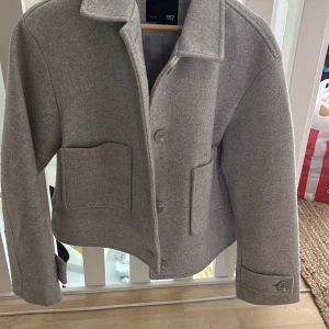 Grå kappa - Stilren kappa från 157 med klassisk krage, stora fickor framtill och knäppning med knappar. Jackan har lång ärm med knappdetalj vid ärmslut. Perfekt för lager-på-lager under höst och vår.