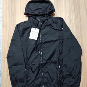 Svart vindjacka från Moncler - Svart vindjacka från Moncler med huva och dragkedja framtill. Jackan har snörning vid halsen och vita, röda och blå detaljer på huvan. Materialet är lätt och syntetiskt, perfekt för blåsiga dagar. Moncler-logga på ärmen och praktiska fickor med knappar.