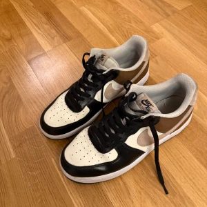 Nike Air Force 1 sneakers svart/vit/brun (ÄKTA) - Nike Air Force 1 sneakers i en clean färgkombo med svart, vit och brun. Klassisk låg modell med perforerad tå, svart snörning och vit sula. Swoosh-loggan i beige mocka på sidan och bruna paneler ger extra stilpoäng. Perfekt för dig som gillar streetwear och tidlös design.