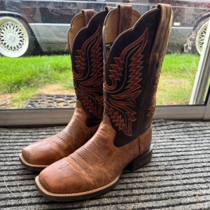 Bruna cowboy boots i skinn - Snygga cowboy boots från Ariat i brunt skinn med mörkbrun skaft och broderade detaljer i rött och beige. Klassisk fyrkantig tå och robust klack. Nypris 2759 säljes för 2199, använda max 3 gånger.