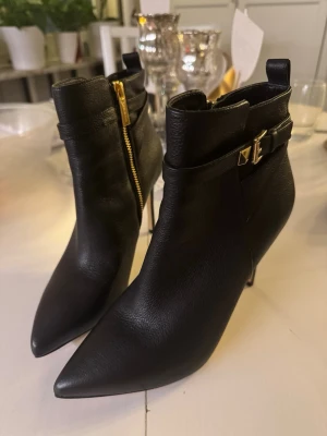Svarta stövletter med spetsig tå och klack - Säljer ett par svarta stövletter i skinn med spetsig tå och hög klack. Skorna har guldfärgade detaljer som dragkedja och spänne vid ankeln. 