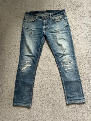 Nudie Jeans blå slitna jeans 34/34 - Säljer ett par blå jeans från Nudie Jeans med slitningar och distressed detaljer på både fram- och baksida. Klassisk femficksmodell med orange sömmar och Nudie-logga på bakfickan. Jeansen har rak passform och är tillverkade i bomull.