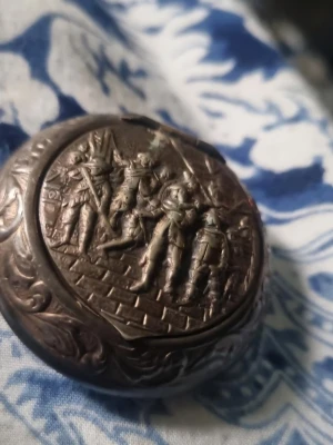 Antik rund snusdosa i silver - Rund snusdosa i silverfärgad metall med detaljerade gravyrer av soldater på locket. Dosan har ett gångjärnslock och vackra ornament längs kanterna. Perfekt för dig som gillar historiska eller unika accessoarer.        Gör en gooogle bildsökning på den 😉