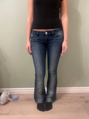 Mörkblå bootcut jeans med låg midja - Säljer ett par mörkblå bootcut jeans med snygg tvätt och låg midja. Jeansen har klassiska fem fickor, markerade sömmar och lätt utsvängda ben. Materialet är stretchigt jeans för skön passform och de har en silverfärgad knapp framtill.
