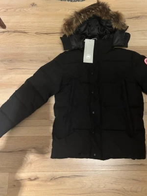 Svart dunjacka från Canada Goose - Svart dunjacka från Canada Goose med fet pälskant på huvan och klassisk logga på ärmen. Jackan har lång ärm, flera fickor och stängs med både dragkedja och knappar. Perfekt för kalla vinterdagar och har en riktigt schysst look.