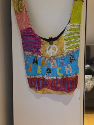 Färgglad axelväska Gandia Beach - Unik axelväska i boho-stil med texten 'Gandia Beach' framtill. Väskan har flera färger som gul, blå, grön, röd och lila, samt dekorativa detaljer som slitsade tygpaneler och en peace-symbol. Tillverkad i bomull med innerfack och knappstängning.