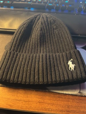 Svart ribbstickad mössa från Polo Ralph Lauren - Svart ribbstickad mössa från Polo Ralph Lauren med klassisk broderad logga framtill. Tillverkad i en mix av bomull och syntetmaterial för både värme och komfort. Perfekt accessoar för kyliga dagar och enkel att matcha med olika outfits.