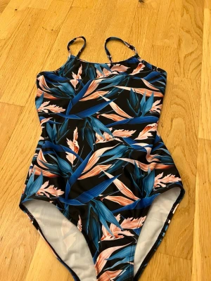 Blommig baddräkt från Ellos - Snygg baddräkt från Ellos med tropiskt blommönster i blått, svart och persikofärgat. Smala axelband och bekväm passform, perfekt för strand eller pool. 