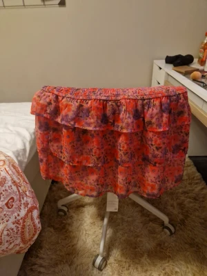 Blommig volangkjol från Bikbok - Säljer en kort kjol från Bikbok i storlek M med blommigt mönster i rosa, lila och rött. Kjolen har bred smock i midjan och flera lager med volanger som ger en flowig och söt look. Perfekt för dig som gillar färgglada och lekfulla plagg.