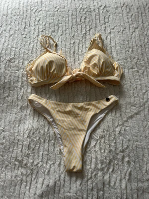 Gulvit randig bikini från Panos Emporio - Säljer en bikini från Panos Emporio i fräscha gula och vita ränder. Bikinin har en knyt-detalj framtill på toppen och justerbara axelband. Både topp och underdel har små guldfärgade logodetaljer. Materialet är mjukt och stretchigt, perfekt för sommaren. Under del storlek S 36 och överdel storlek 40 L 