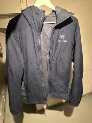 Arcteryx jacka M - Säljer en mörkblå vindjacka från Arc'teryx med huva och dragkedja framtill. Jackan är lätt och smidig, perfekt för blåsiga dagar. Diskret logga på bröstet och enkel design utan onödiga detaljer. Passar dig som gillar funktion och stil.