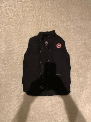 Svart dunväst från Canada Goose - Säljer en svart dunväst från Canada Goose med klassisk logga på bröstet. Västen har hög krage, dragkedja och knappar framtill samt praktiska fickor. Perfekt för lager-på-lager och riktigt snygg till höst- och vinteroutfits.