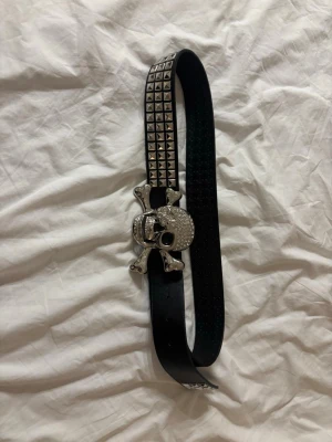 Svart bälte med dödskalle och nitar - Säljer ett svart bälte med stora silvriga nitar och ett riktigt maffigt dödskalle-spänne dekorerat med glittrande stenar. Jag är lite osäker på storleken men generellt sätt så bär jag mellan XXS-M.