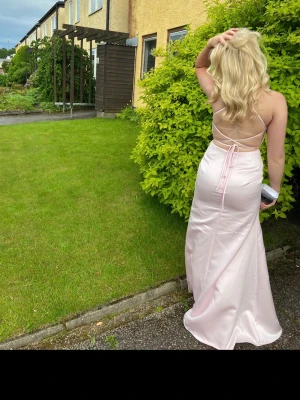 Ljusrosa balklänning  - Ljusrosa balklänning från JJs House. Glansigt material med v-ringning, tunna axelband, en slits vid benet och öppen rygg med knytning. Figurnära passform med draperade detaljer. Använd vid ett tillfälle, i fint skick. Perfekt till bal. Storlek 32 men man kan dra åt den själv i ryggen.