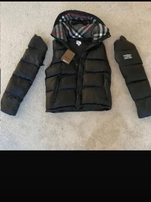 Svart pufferjacka från Burberry - Säljer en svart pufferjacka från Burberry med avtagbara ärmar och klassiskt rutigt foder i huvan. Jackan har dragkedja och knappar framtill, två sidofickor med dragkedja och Burberry-logga på ena ärmen. Perfekt för kalla dagar och kan bäras både som jacka och väst.