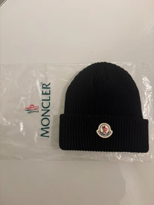 Svart Moncler mössa i ull - Snygg svart ribbstickad mössa från Moncler med klassisk logotyp framtill. Mössan är tillverkad i mjuk ull och har en bred uppvikt kant som ger en cool och tidlös look. Perfekt för dig som vill ha både stil och värme under kyliga dagar.