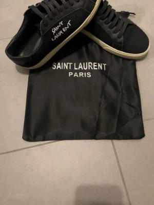 Svarta Saint Laurent sneakers - Säljer ett par svarta Saint Laurent sneakers med vit text på sidan och klassisk vit sula. Skorna har snörning och är tillverkade i en mix av skinn och mocka för en lyxig känsla. Medföljer original dustbag från Saint Laurent Paris.