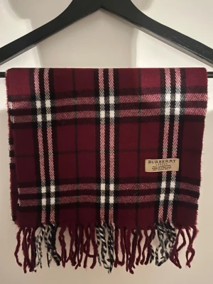 Burberry halsduk - Säljer en klassisk vinröd halsduk från Burberry i 100% lammull. Halsduken har det ikoniska rutiga mönstret i vinrött, svart och vitt samt fransar i ändarna. Perfekt accessoar för att lyfta din outfit med en tidlös brittisk vibe.
