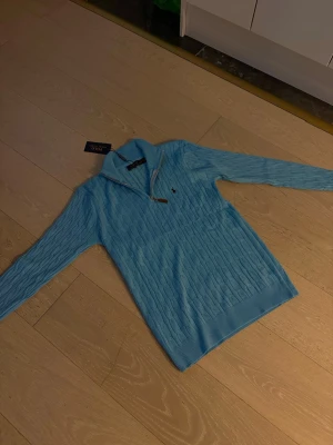 Ljusblå kabelstickad tröja Polo Ralph Lauren - Supersnygg ljusblå kabelstickad tröja från Polo Ralph Lauren med halv dragkedja och klassisk logga på bröstet. Tröjan har lång ärm, ribbade muddar och är perfekt för dig som vill ha en clean och stilren look. Fått den som present värt 2000kr men är inte i behov av den.