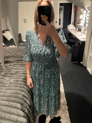 Blommig långklänning med v-ringning - Säljer en grön långklänning med blommigt mönster och v-ringad omlottfront. Klänningen har korta puffärmar och ett smockat midjeparti som ger en snygg siluett. Perfekt för dig som gillar romantiska och färgglada plagg.