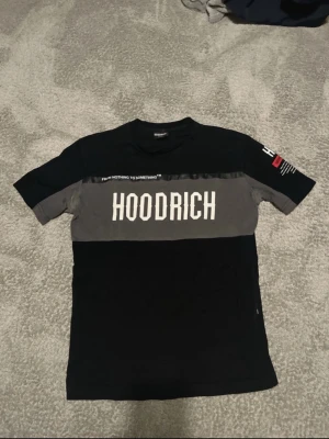 Svart och grå HOODRICH t-shirt - Snygg t-shirt från HOODRICH i svart och grå med stor vit logga över bröstet och textdetaljer på ena ärmen. T-shirten har klassisk passform och är gjord i mjuk bomull. Perfekt för dig som gillar streetwear och vill sticka ut.